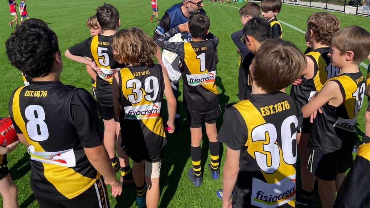 Junior football team wearing black and yellow fisiocrem-sponsored jerseys gathered around a coach.