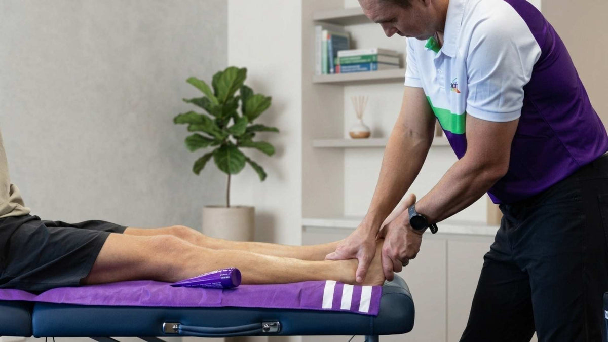 Practitioner holding a client's foot and ankle on a treatment table beside a purple fisiocrem tube.