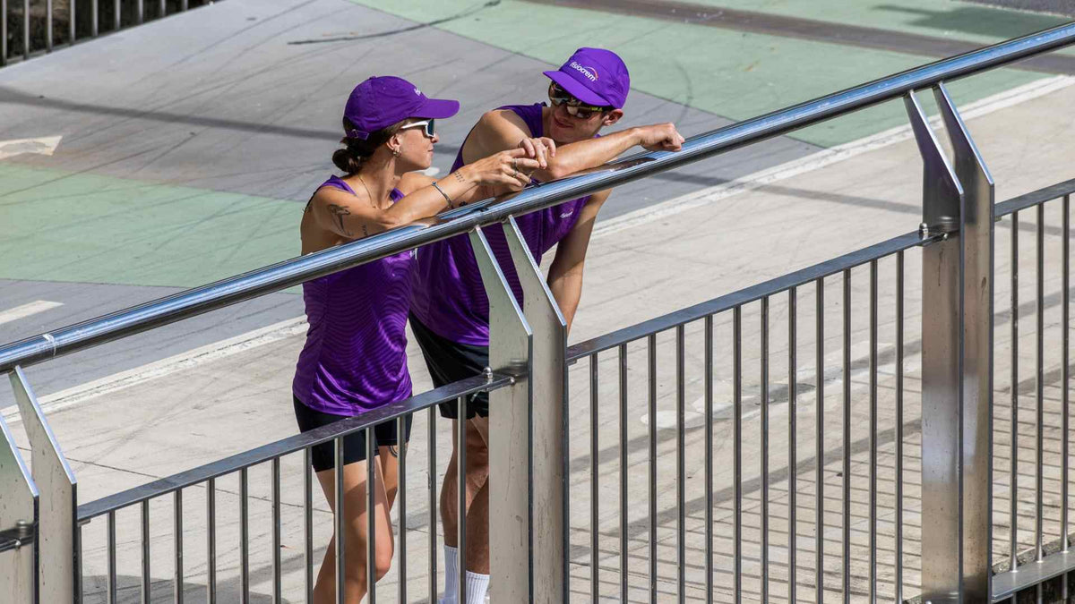 Two people in purple running tops and caps leaning on a metal railing.