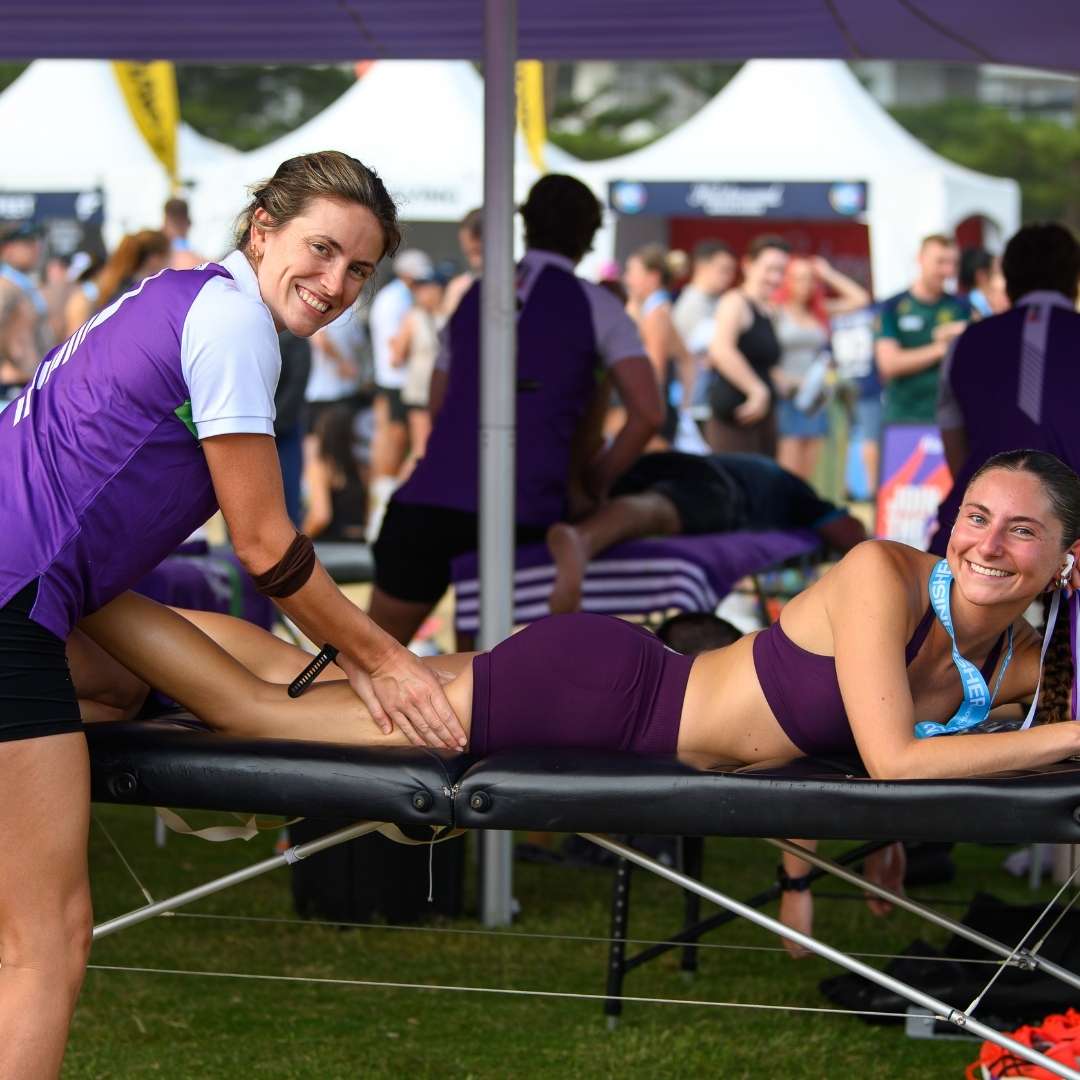 Smiling practitioner massaging a runner's leg on a treatment table beneath an event marquee.