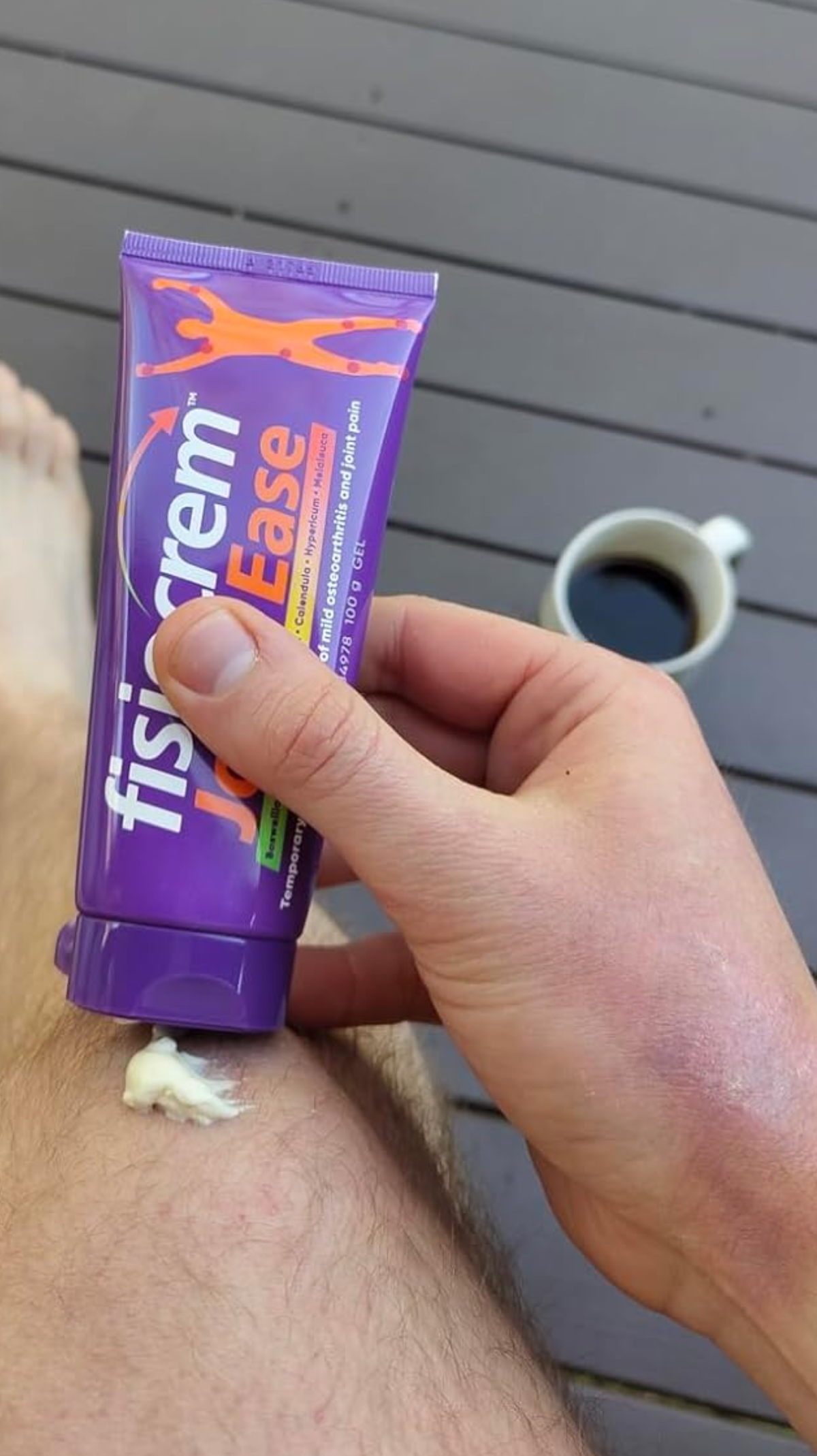 Hand holding a fisiocrem Joint Ease joint pain relief cream tube beside cream on a knee and a cup of coffee.