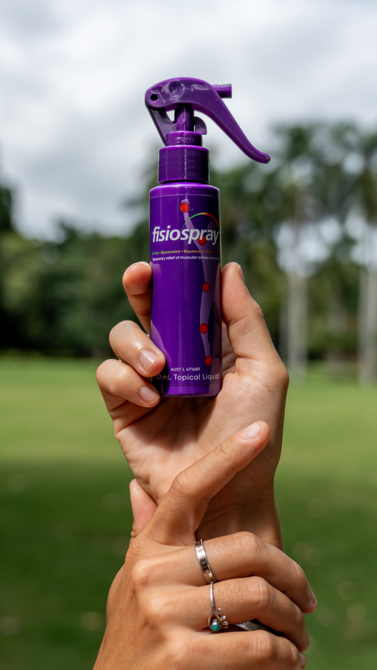 Hand holding a purple fisiospray nerve pain relief spray bottle upright against a grassy park background.